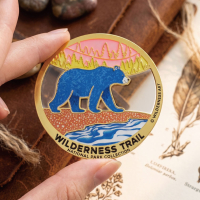 Hand holding a round custom metal souvenir featuring a blue bear, translucent enamel forest details, and full-color UV-printed water artwork.