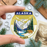 Hand holding an Alaska-themed custom metal souvenir with mountain scenery, evergreen trees, a bear illustration, and transparent imitation hard enamel details.